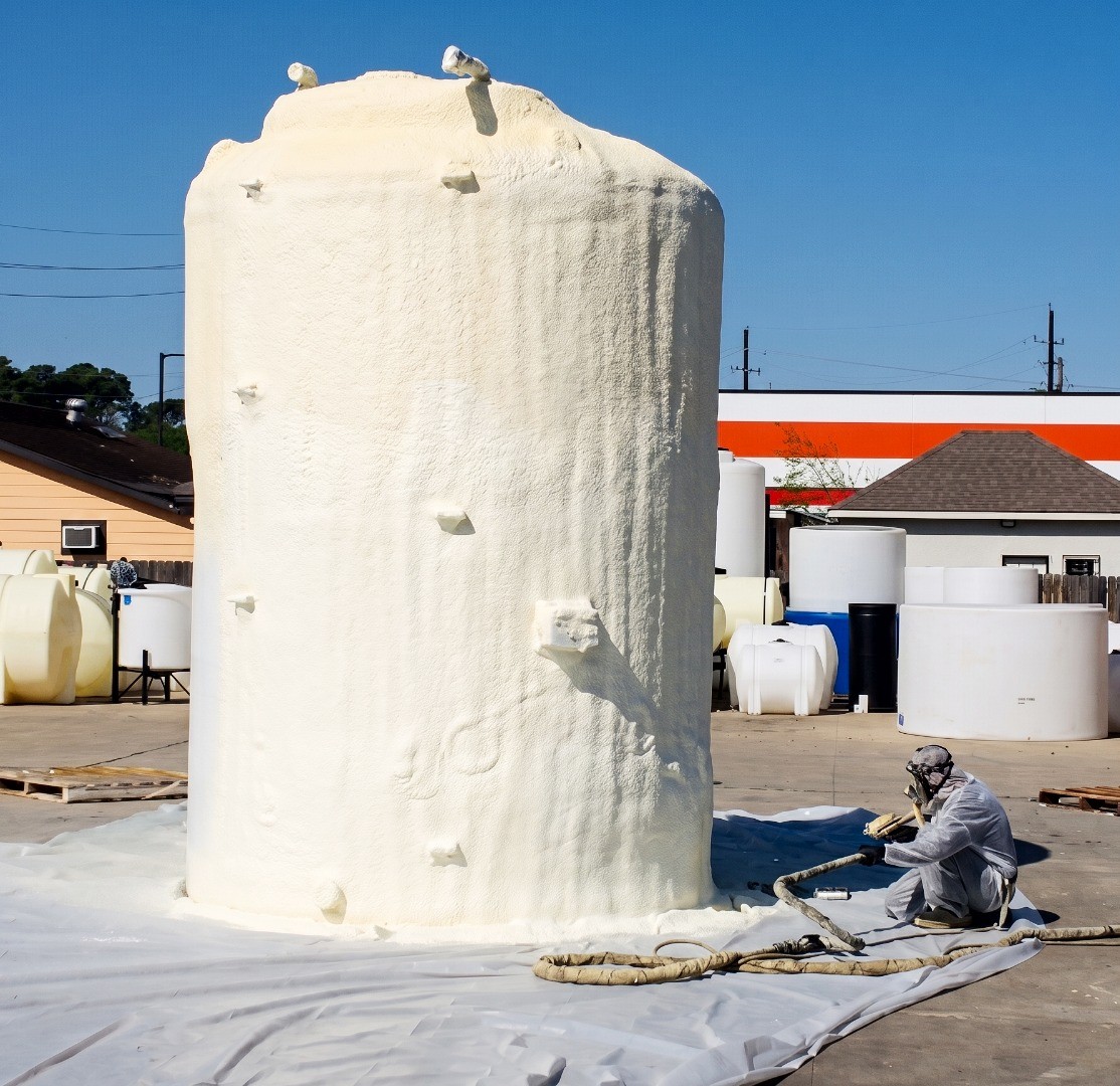 Worker in full white protective suit and respirator applying spray foam insulation to large vertical storage tank