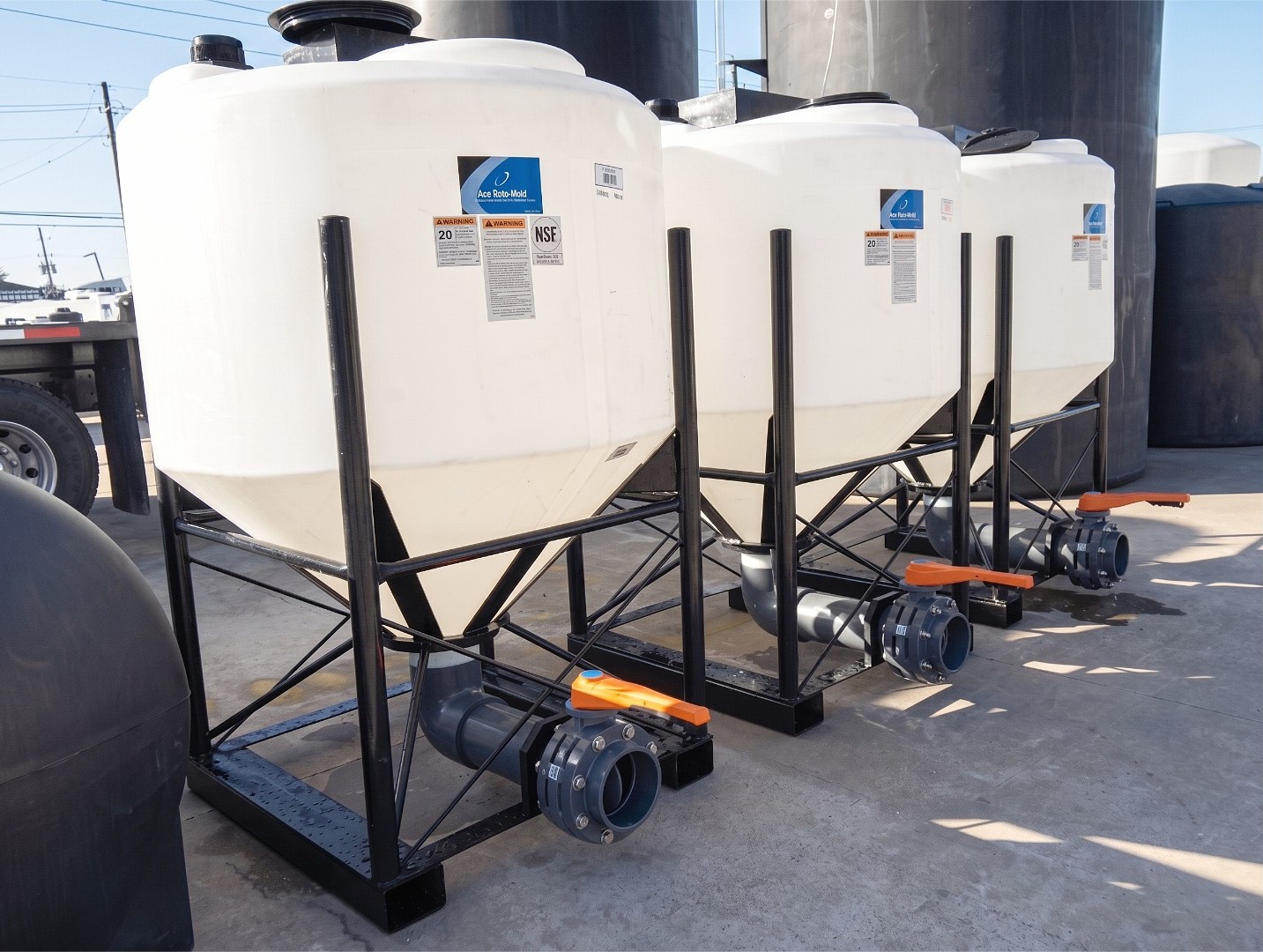A row of three white cone bottom poly tanks on black steel elevated stands with outlet valves and fittings