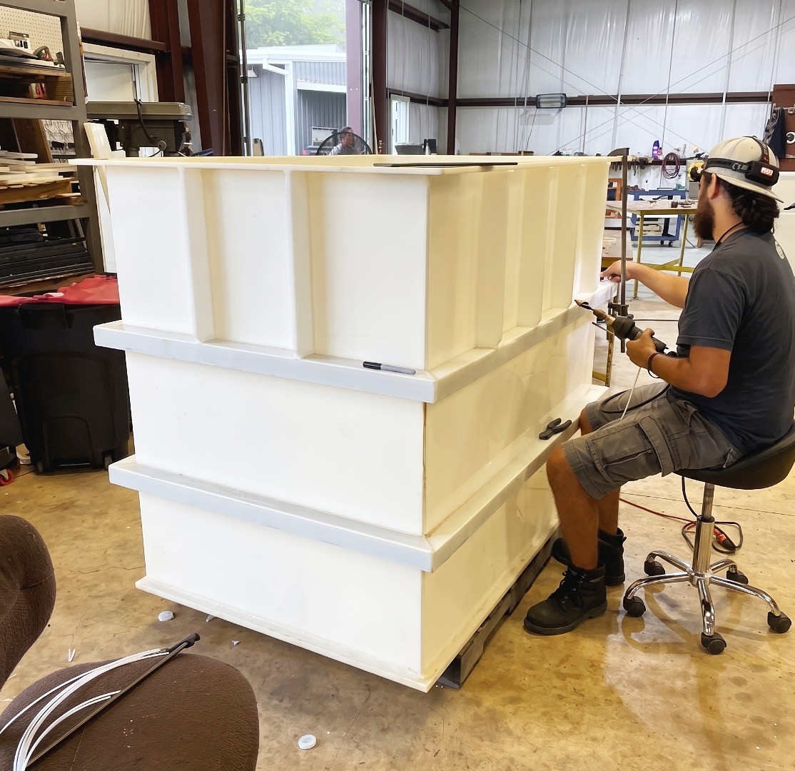 A workshop technician using a plastic welding tool to seal seams on a large, custom-made white rectangular tank