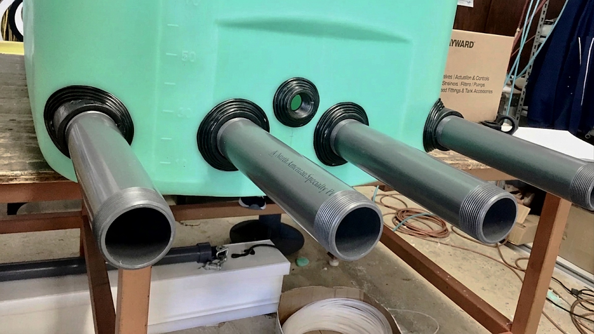 Close-up of four grey threaded PVC pipes protruding through black bulkhead fittings on a light green industrial tank
