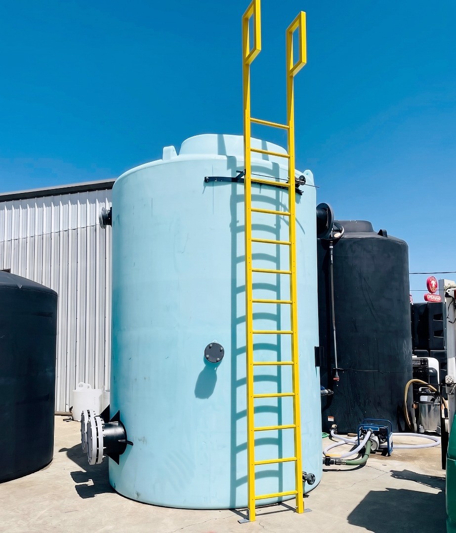 A tall, light blue vertical storage tank with yellow safety ladder in an outdoor industrial storage area