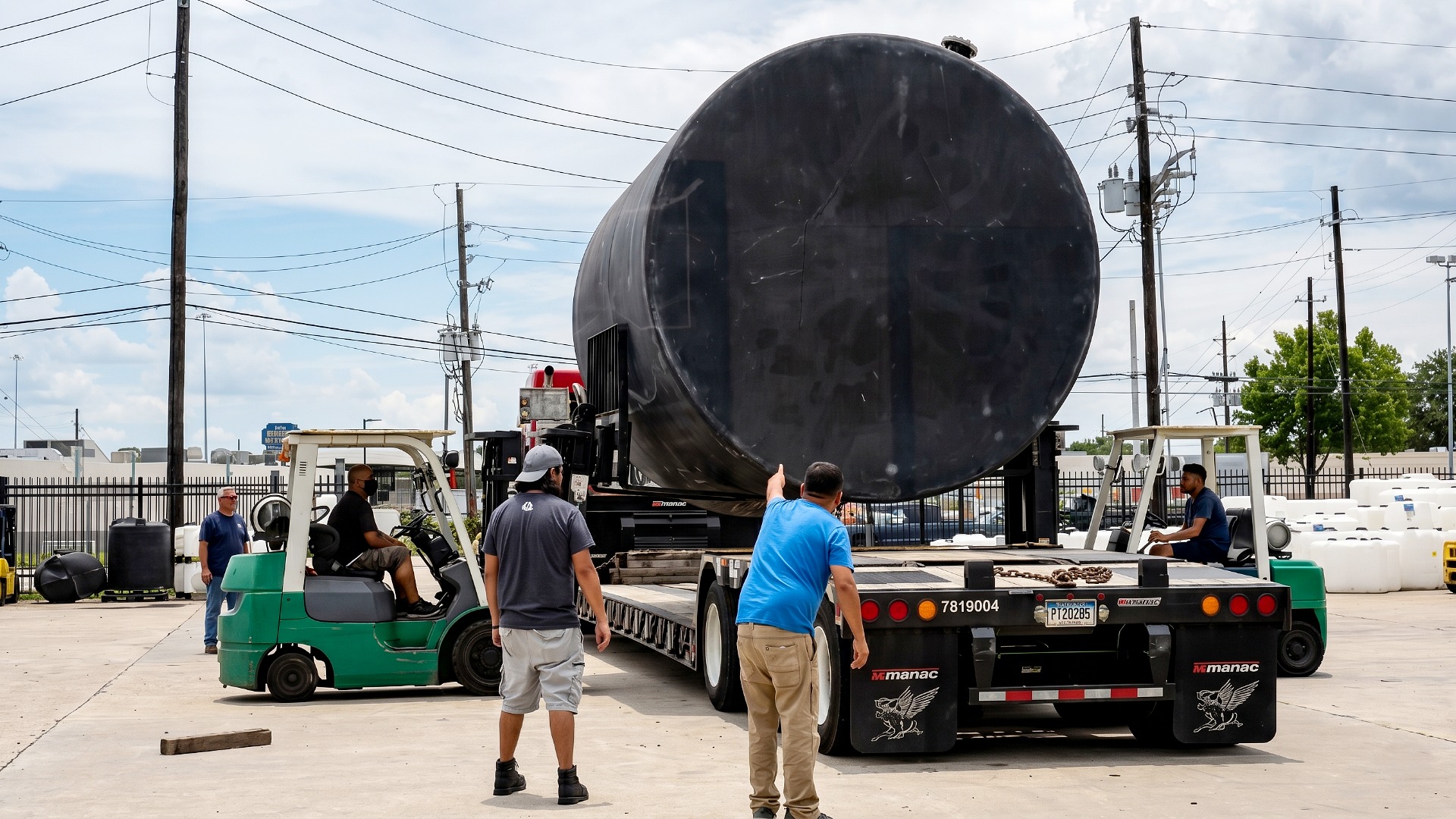 A massive black chemical storage tank being loaded onto a flatbed trailer using forklifts and a team of workers