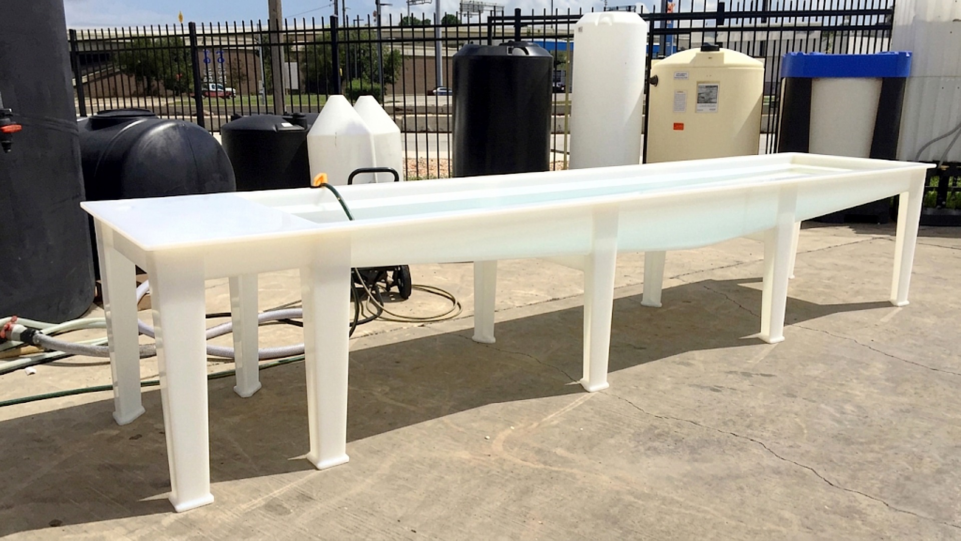 An exceptionally long and narrow white plastic trough sink supported by six integrated legs, shown in an outdoor staging area