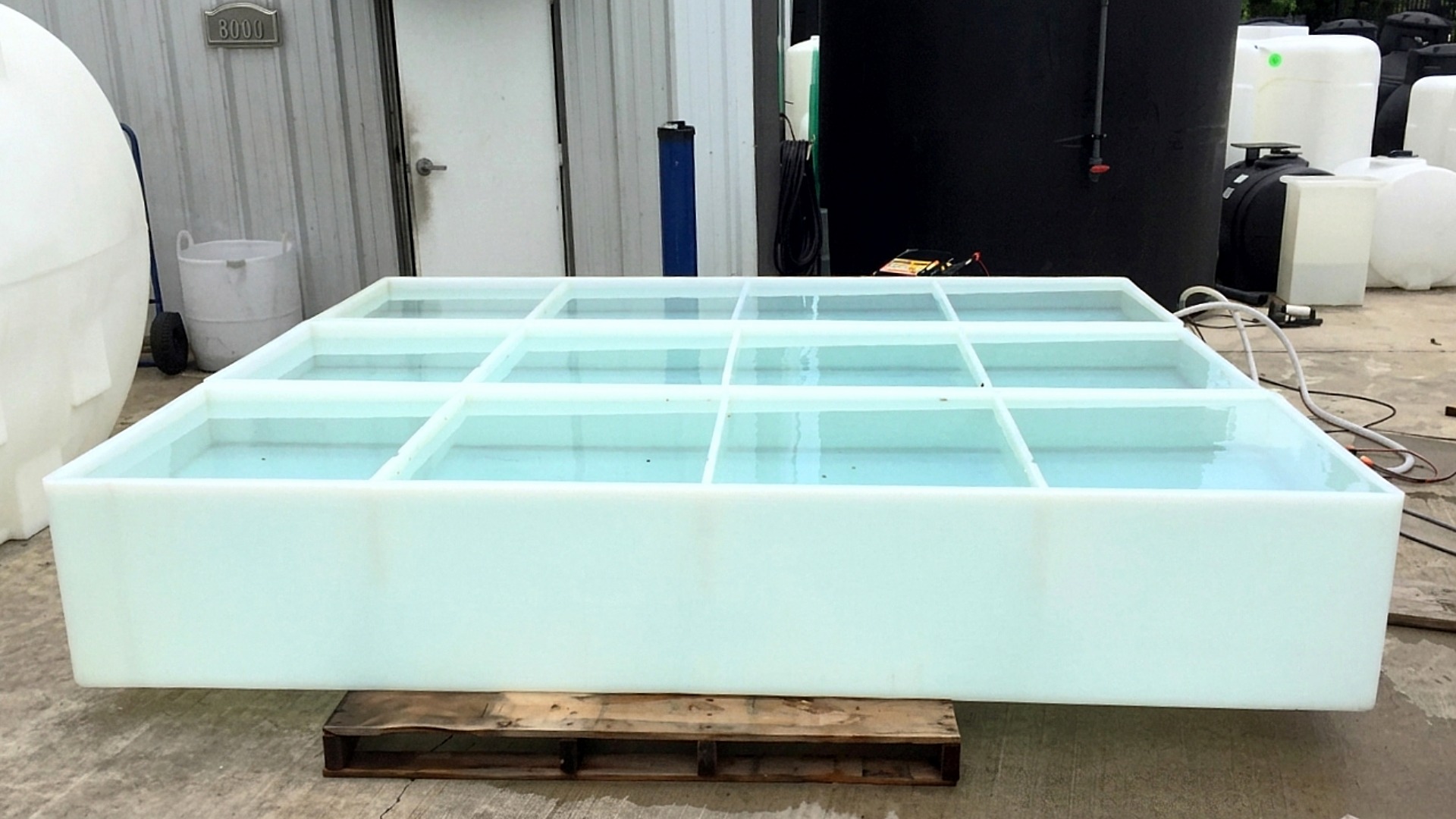A low profile white rectangular tank divided into a nine-compartment grid, filled with water and resting on a wooden pallet