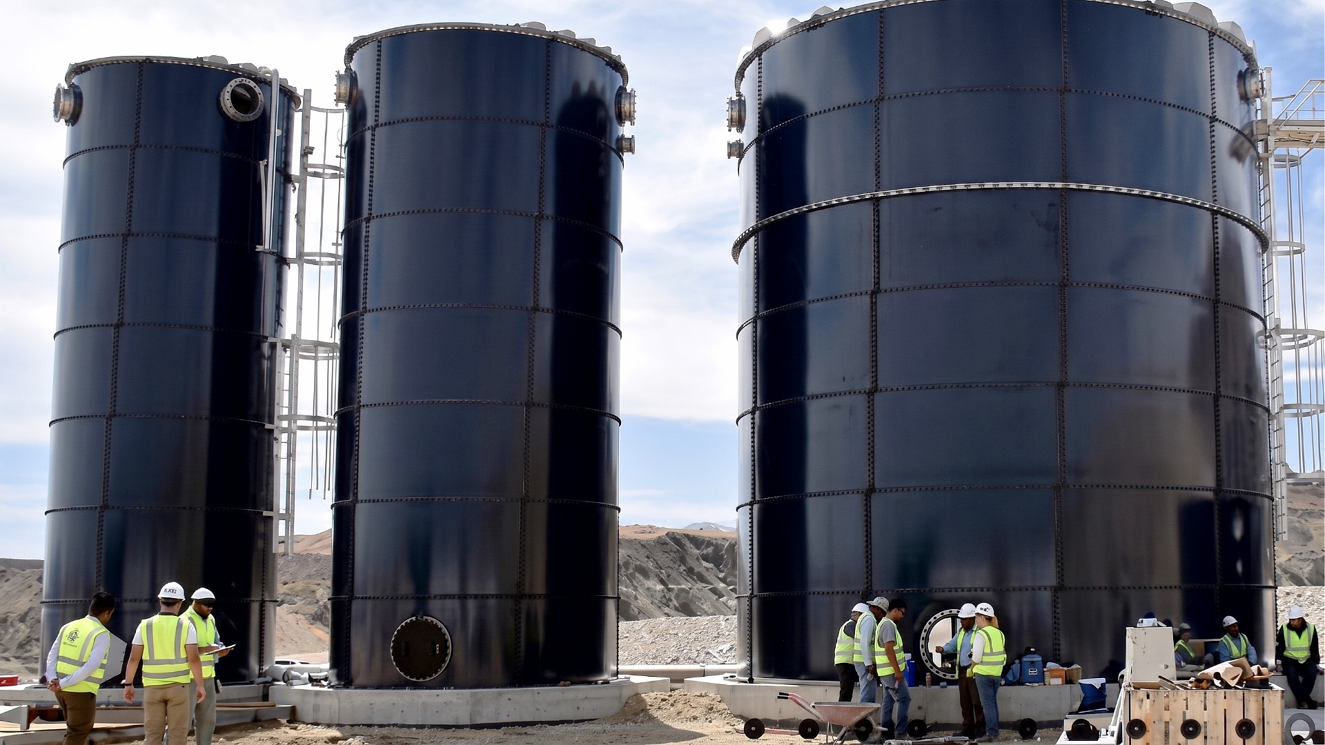 Three tall, large capacity bolted steel storage tanks being installed at a data center construction site. Construction workers are seen in front