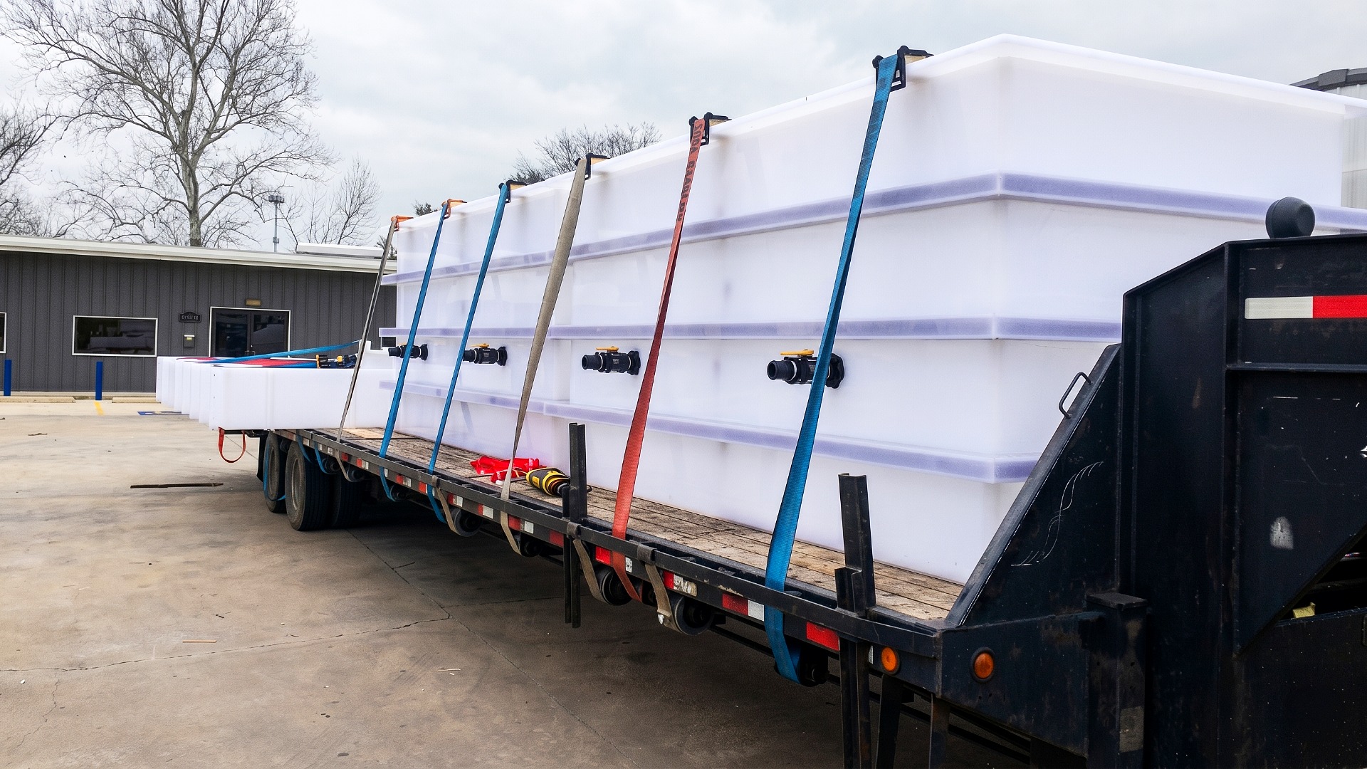 A row of large custom-built polypropylene containment tanks secured with heavy-duty straps on a flatbed trailer for delivery